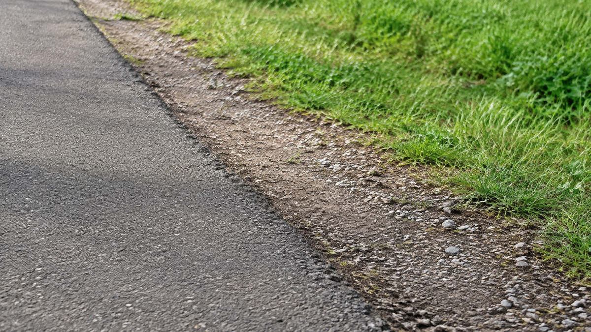 Asphalt grenzt an einen Streifen aus Erde und kleinen Steinen mit Gras dahinter